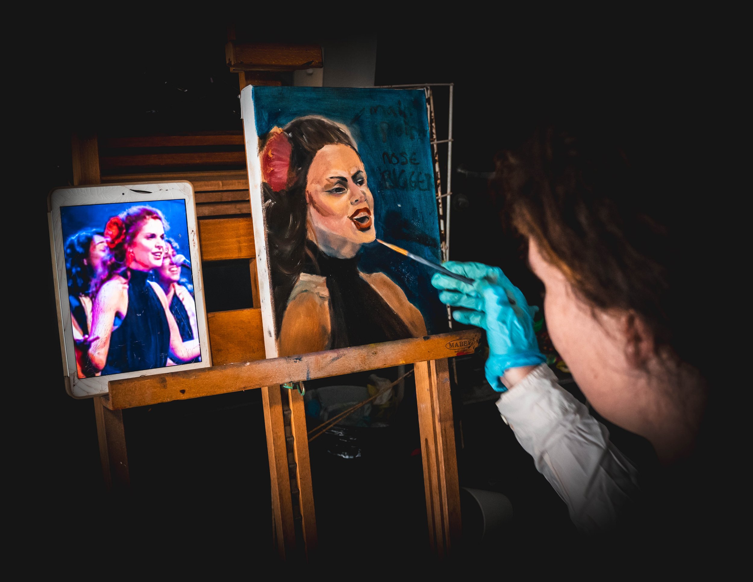 Artist painting portrait on easel, referencing image from tablet.