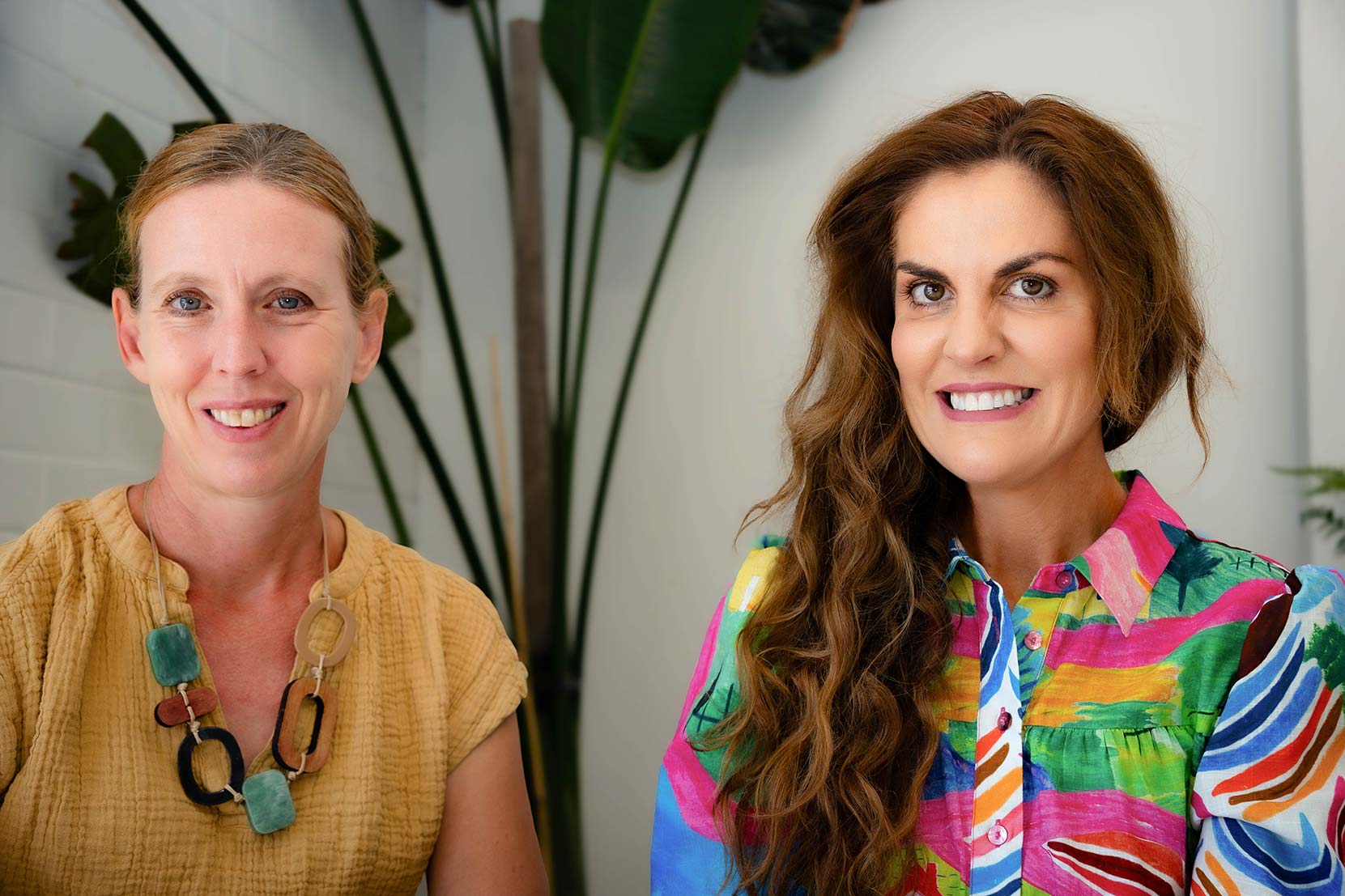 Two women smiling, one in mustard, one in multi-coloured shirt.